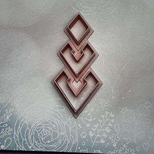 May include: Set of three pink metal cookie cutters shaped like a diamond with a heart inside. The cutters are stacked on top of each other, with the largest cutter at the bottom and the smallest cutter at the top.