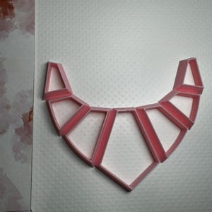 Puede incluir: Un collar de plástico rosa hecho de múltiples formas geométricas. El collar está hecho de 10 piezas que están conectadas entre sí para formar una forma curva.