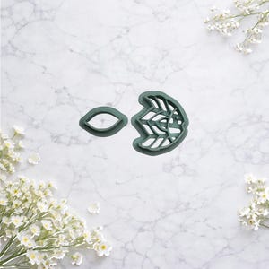 Puede incluir: Dos cortadores de galletas de plástico verde. Uno tiene forma ovalada y el otro un diseño floral. Los cortadores están sobre una superficie de mármol blanco con flores blancas en las esquinas.