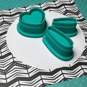 May include: Three teal plastic cookie cutters: a heart shape, a long oval, and a shorter oval. The cutters are on a white background with a black and white chevron pattern.