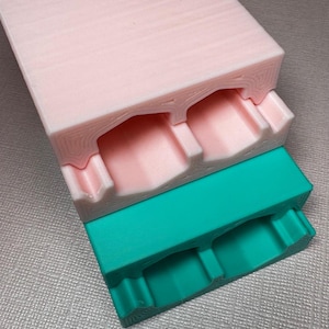 May include: Two stacked 3D printed molds, one pink and one teal, with two rectangular cavities in each. The molds are designed for casting resin.