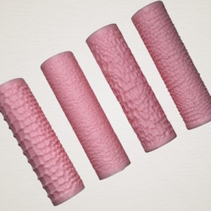 Può includere: Quattro cilindri rosa con superfici strutturate disposti diagonalmente. Gli oggetti hanno una tonalità rosa uniforme e una consistenza leggermente ruvida e irregolare, suggerendo un materiale poroso. I cilindri sono distanziati uniformemente.