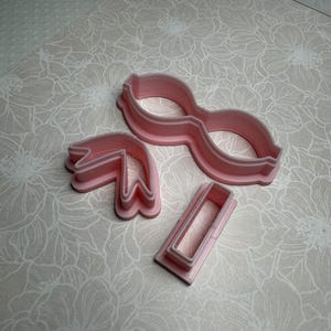 Pode incluir: Um conjunto de três cortadores de biscoitos de plástico rosa. Os cortadores têm a forma de óculos, um laço e um retângulo.