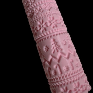 Può includere: Un mattarello rosa con motivi in rilievo dettagliati. Il design include forme floreali, a cuore e geometriche, oltre a motivi di alberi di Natale e bastoncini di zucchero. Il mattarello è realizzato in un materiale rosa chiaro.
