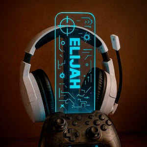 May include: A gaming setup with a black controller, white headphones, and a glowing blue acrylic panel with the name "ELIJAH" and the text "DESIGN 4". The headphones are resting on the panel and controller.