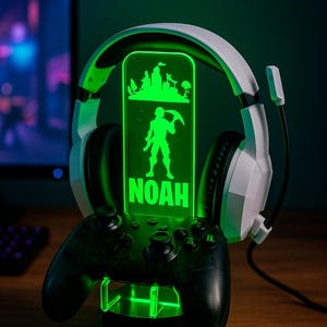 Peut inclure: Un support pour casque et manette de jeu avec un design vert illuminé. Le support présente une silhouette de personnage avec le nom "NOAH" et le texte "DESIGN 3". Le casque est blanc et noir.