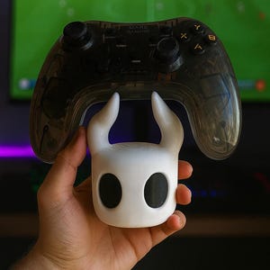 May include: A white and black video game controller accessory shaped like a character's head, held beneath a transparent black controller. The controller has the text "MARS GAMING" on it. The background is a blurred image of a television screen.