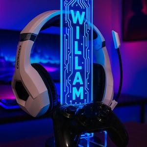 Può includere: Un supporto per cuffie da gioco con un pannello luminoso a LED blu che mostra il nome "WILLIAM" in bianco. Il supporto contiene una cuffia bianca e nera e un controller di gioco nero. Il design è etichettato "DESIGN 1".
