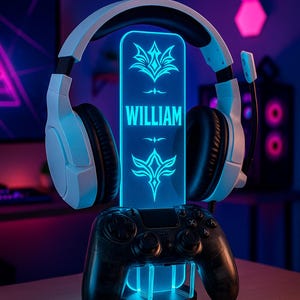 Könnte beinhalten: Ein Gaming-Headset liegt auf einem schwarzen Controller-Ständer mit blauer LED-Beleuchtung. Eine blaue Acrylplatte zeigt den Namen "WILLIAM" und dekorative Embleme. Auf der Basis des Ständers steht "DESIGN 2".