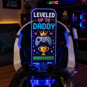 May include: A gaming-themed night light with the text "LEVELED UP TO DADDY" in pixelated font. The design includes a controller, crown, and trophy. The light is blue and is set on a black controller stand with a white headset.