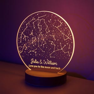 Peut inclure: Une veilleuse personnalisée avec un motif de constellation. Le disque en acrylique transparent affiche une carte des étoiles avec les noms "Julia & William" et la phrase "Love you to the moon and back". Elle repose sur une base en bois.