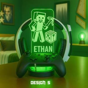 Puede incluir: Lámpara LED verde brillante con un personaje pixelado sosteniendo un pico y un cubo, con el nombre "ETHAN" debajo. Un mando de juego negro se encuentra en la base, con unos auriculares detrás de la lámpara. El texto "DESIGN 5" está en la parte inferior.