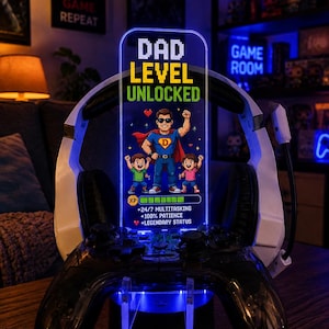 May include: A gaming-themed LED light with the text "DAD LEVEL UNLOCKED" and a pixelated superhero graphic. The light is mounted on a black game controller stand, with a white and black headset resting on top. The design includes the text "DESIGN 6".
