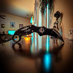 May include: A black karambit knife with a silver blade and a skull design on the handle. The knife has the word "BANDERZA" engraved on the handle.