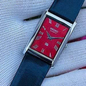 Könnte beinhalten: Eine rechteckige Seiko Quartz Armbanduhr mit einem leuchtend roten, strukturierten Zifferblatt und silberfarbenen römischen Ziffern als Stundenmarkierungen. Die Uhr hat ein poliertes silberfarbenes Metallgehäuse und ein glattes schwarzes Lederarmband.