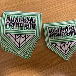 May include: Green and white embroidered patches with the text "Dimebag Darrell" and "Army". The patches have a shield shape with a white triangle at the bottom. Several patches are stacked together.