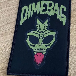 Pode incluir: Remendo de tecido preto com o texto "Dimebag" em verde e uma criatura alienígena verde com uma língua vermelha.