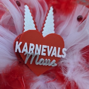 May include: A vibrant red and white feathered accessory with a red heart-shaped centerpiece. The heart features the text "KARNEVALS Maus" in white and silver lettering, and is adorned with white, jagged-edged accents.