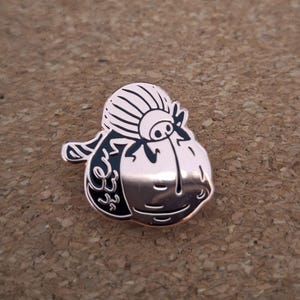 May include: A rose gold metal enamel pin shaped like a stylized sheep with a black and white face and a textured woolly top. The design features a smooth metallic finish on the lower portion and detailed black enamel lines defining the features.