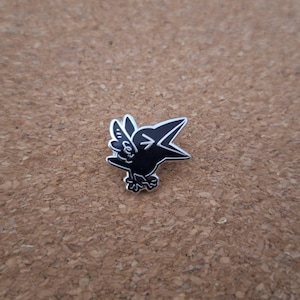 Könnte beinhalten: Eine schwarz-weiße Emaille-Anstecknadel, die eine stilisierte Cartoon-Krähe mit weit geöffnetem Schnabel zeigt. Das Design verwendet eine kräftige schwarze Füllung mit weißen Umrandungen, um die Flügel, Füße und Gesichtszüge des Vogels zu definieren.