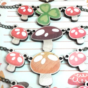 May include: A set of five necklaces featuring wooden charms with mushroom, acorn, and clover designs. The charms are painted in various shades of red, green, brown, and white.