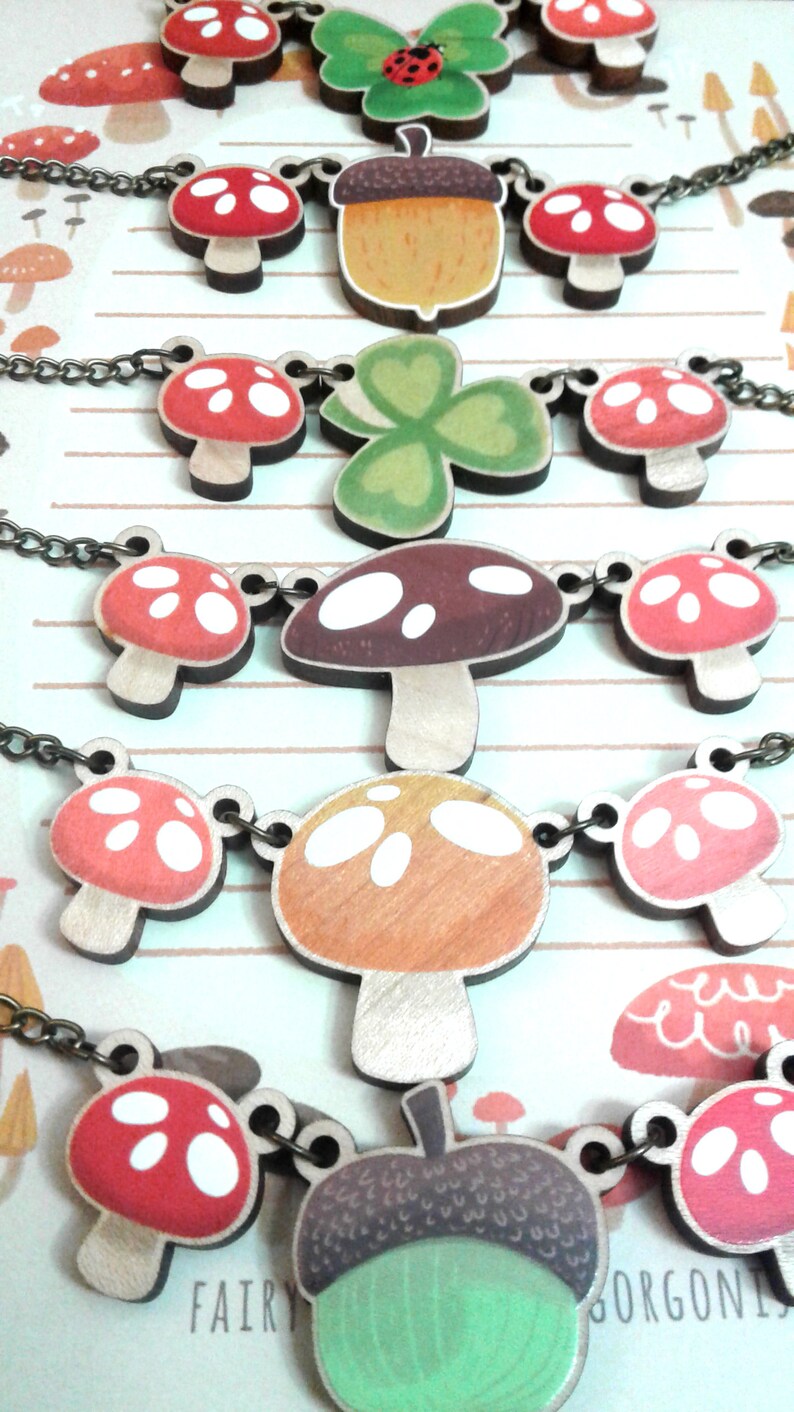 May include: A set of five necklaces featuring wooden charms with mushroom, acorn, and clover designs. The charms are painted in various shades of red, green, brown, and white.