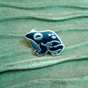 May include: A black and white enamel pin featuring a frog with white spots. The frog is in a sitting position with its legs tucked under its body.