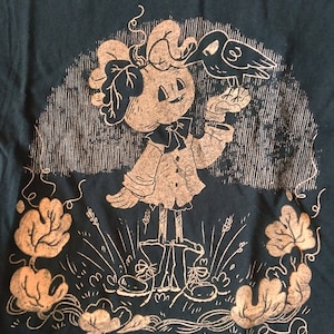 May include: A black t-shirt with a gold print of a pumpkin-like character wearing a bow tie and holding a bird. The character is standing in a field of grass and leaves.