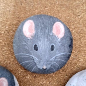 May include: A circular pinback button featuring a detailed illustration of a grey rat face. The rat has prominent pink ears, dark black eyes, a small pink nose, and long white whiskers extending across the dark grey fur texture of the design.