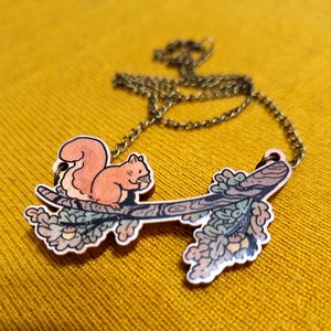 Op de afbeelding: Een houten ketting met een eekhoorn die op een tak zit met bladeren en eikels. De ketting is gemaakt van geschilderd hout en heeft een bronzen ketting.
