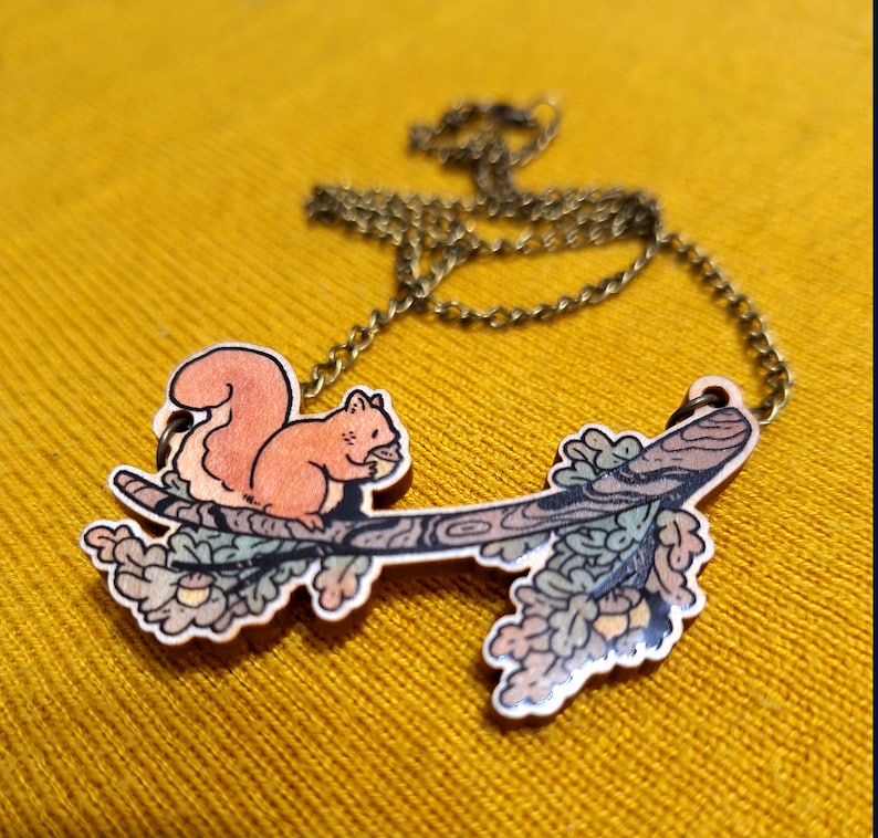 Op de afbeelding: Een houten ketting met een eekhoorn die op een tak zit met bladeren en eikels. De ketting is gemaakt van geschilderd hout en heeft een bronzen ketting.