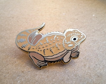 Op de afbeelding: Een gouden emaille pin met een cartoon baardagame met een lichtbruin lichaam en een witte buik. De draak heeft een stekelige rug en een groot, rond oog.