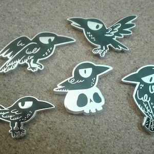 May include: Five black and white enamel pins featuring different crow designs. One crow is perched on a skull, another is flying, and the rest are standing.