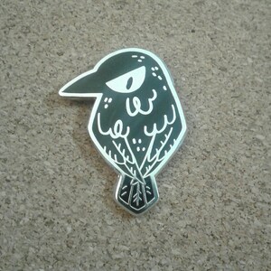 May include: A black and white enamel pin of a cartoon crow with a grumpy expression. The crow is outlined in white and has a black eye with a white pupil.