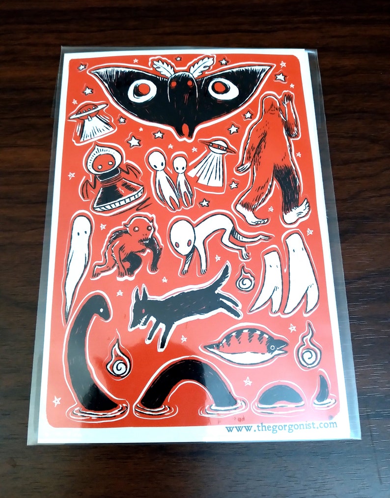 May include: A red sticker sheet with black and white illustrations of cryptids and UFOs. The sheet includes a moth, Bigfoot, aliens, a dog, and other mysterious creatures. The website address www.thegorgonist.com is printed at the bottom.