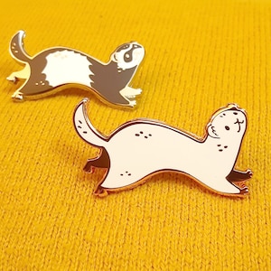 May include: Two gold-coloured ferret pins. One pin is black and white and the other is white. The pins are on a yellow knitted surface.
