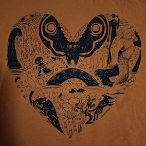May include: Mustard-coloured t-shirt with a navy blue heart-shaped design. The design is filled with cryptids and mythical creatures, including a moth, Bigfoot, aliens, and other fantastical beings.