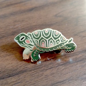 May include: Gold-tone turtle pin with a green shell and pink accents. The turtle is walking with its head and legs extended.