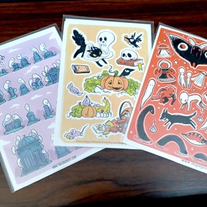 May include: Three sticker sheets with Halloween illustrations. One shows ghosts and gravestones, another has a black cat, pumpkin, and skull, and the third features a moth, aliens, and a dog. Each sheet is in a clear plastic sleeve.