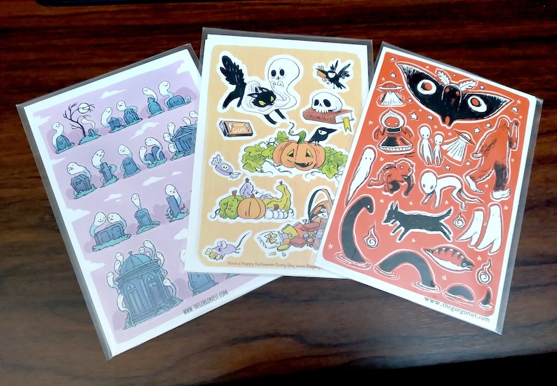 May include: Three sticker sheets with Halloween illustrations. One shows ghosts and gravestones, another has a black cat, pumpkin, and skull, and the third features a moth, aliens, and a dog. Each sheet is in a clear plastic sleeve.