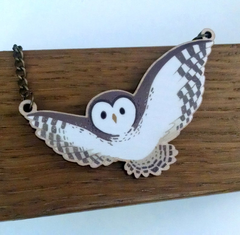 May include: A white owl with brown and white patterned wings, flying with its wings spread wide, is depicted on a wooden pendant necklace.