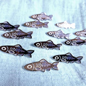 Könnte beinhalten: Eine Sammlung kleiner fischförmiger Emaille-Anstecknadeln in einer dunkelblauen und silbernen Farbpalette. Jede Anstecknadel zeigt ein stilisiertes Skelettfisch-Design mit einem markanten kreisförmigen Auge, detaillierten Flossen und einem metallischen Finish.