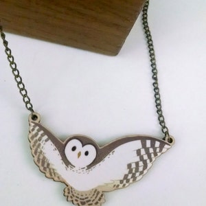 May include: A white and brown owl pendant necklace with a bronze chain. The owl is flying with its wings spread out. The pendant is made of wood and is painted with a realistic design.