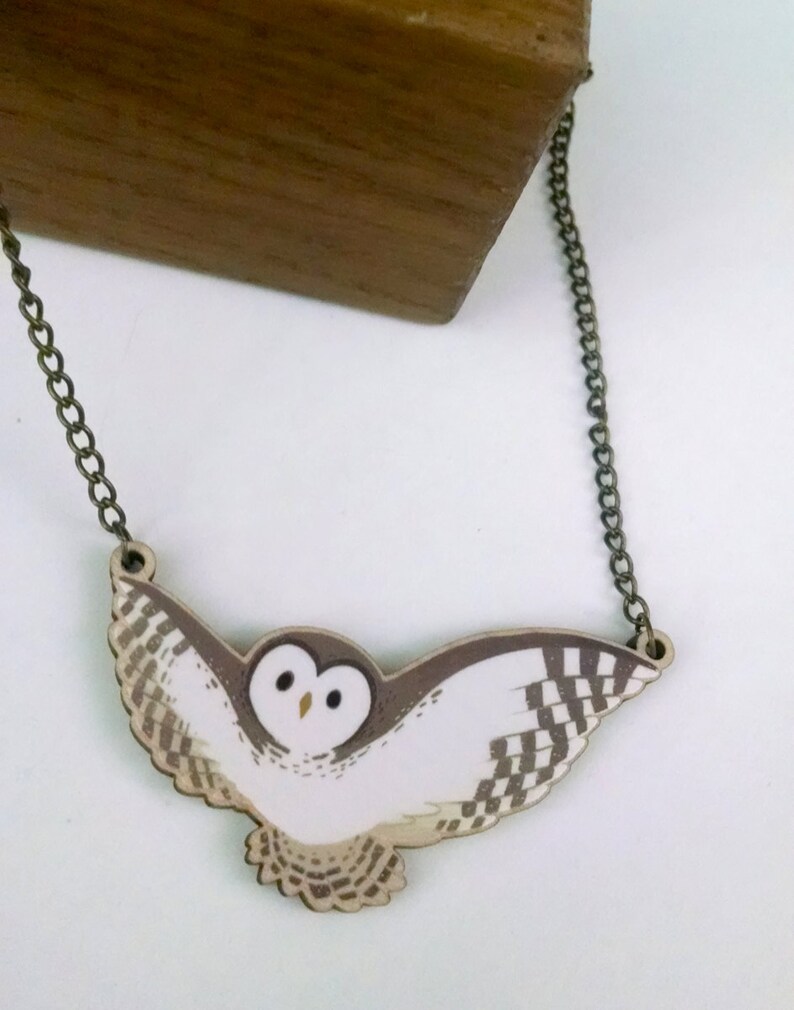 May include: A white and brown owl pendant necklace with a bronze chain. The owl is flying with its wings spread out. The pendant is made of wood and is painted with a realistic design.