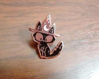 May include: Black enamel pin of a cat wearing a witch hat with a crescent moon and stars. The cat is sitting with its tail curled around its body.