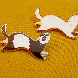 May include: Two gold-coloured enamel pins featuring a cartoon ferret. One ferret is white with black eyes and the other is black with white markings.