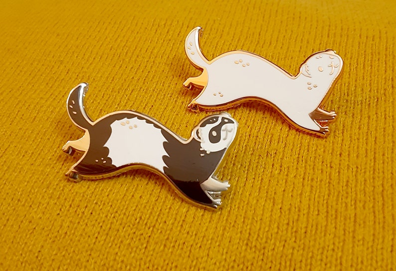 May include: Two gold-coloured enamel pins featuring a cartoon ferret. One ferret is white with black eyes and the other is black with white markings.