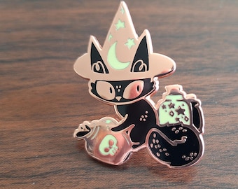 以下が含まれることがあります： 三日月と星がついたローズゴールドの魔女の帽子をかぶった黒い猫。猫は、どくろと星が描かれた2本の魔法薬の瓶を持っています。瓶もローズゴールドです。