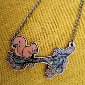 Op de afbeelding: Een houten ketting met een eekhoorn die een noot vasthoudt op een tak met bladeren. De ketting ligt op een gele stoffen achtergrond.