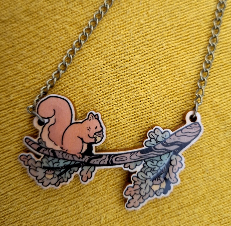 Op de afbeelding: Een houten ketting met een eekhoorn die een noot vasthoudt op een tak met bladeren. De ketting ligt op een gele stoffen achtergrond.
